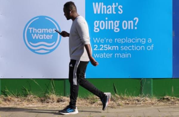 A black man in a long-sleeved T-shirt and track-suit bottoms, wearing earbuds and looking at his mobile phone, passes a Thames Water billboard that says: “What’s going on? We’re replacing a 2.5km section of water main”