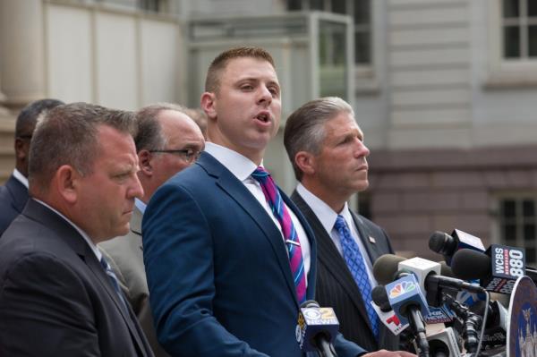 Assemblyman Mike LiPetri speaking at a press co<em></em>nference a<em></em>bout new proposed legislation, surrounded by men in suits and speaking into microphones