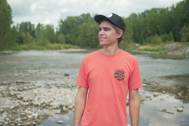 A young man in a red T-shirt and a black ba<em></em>seball cap stands on the rocky shore of a river. 