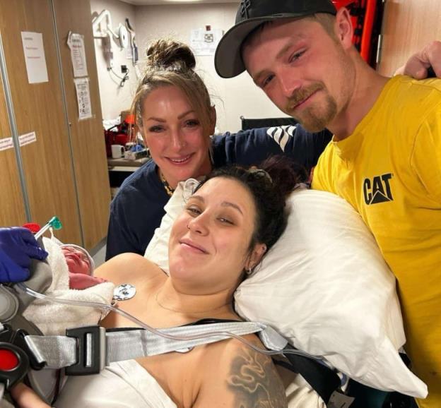 A woman lying on a pillow holds her newborn baby. A smiling woman and man wearing a yellow t-shirt stand behind her.