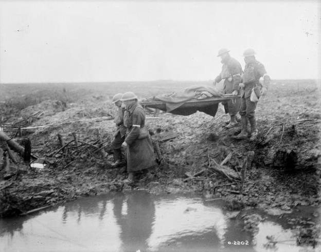 Four soldiers carry a stretcher, bearing a wounded man, across the a barren landscape. 