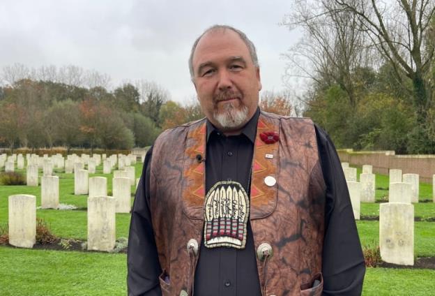 A bearded man, wearing a leather vest, stands in a ceme<em></em>tary. 