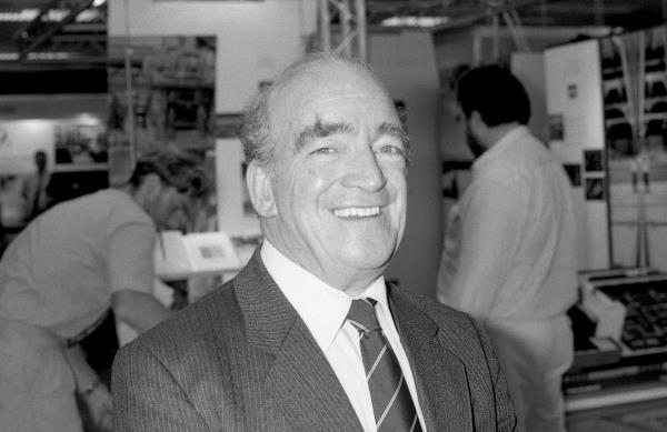 Smiling man in suit and tie, with factory workers behind him.