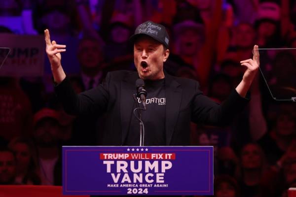 Elon Musk pointing in the air and exclaiming excitedly while standing at a Trump Vance podium at a rally.