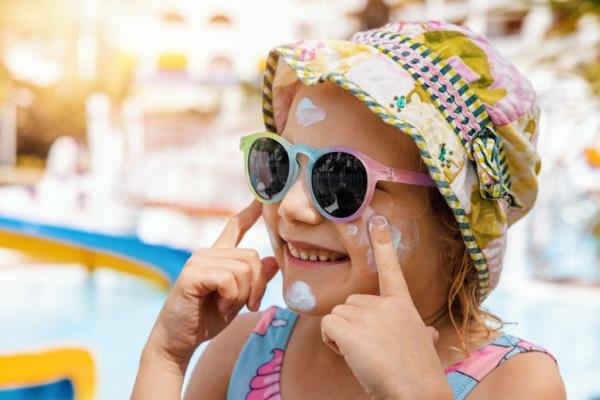 A little girl wearing a hat and sunglasses puts sunscreen on her cheeks.