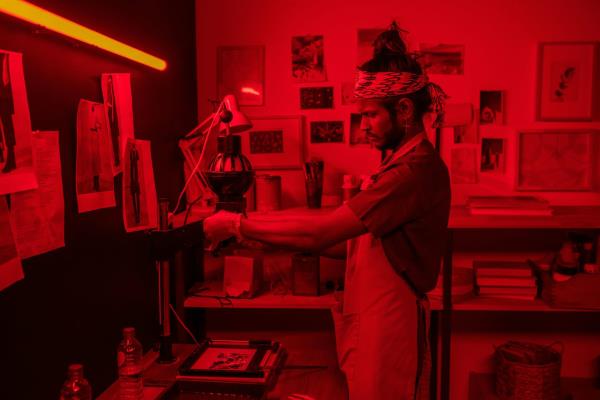 A young male photographer working in the red light of a dark room developing pictures.